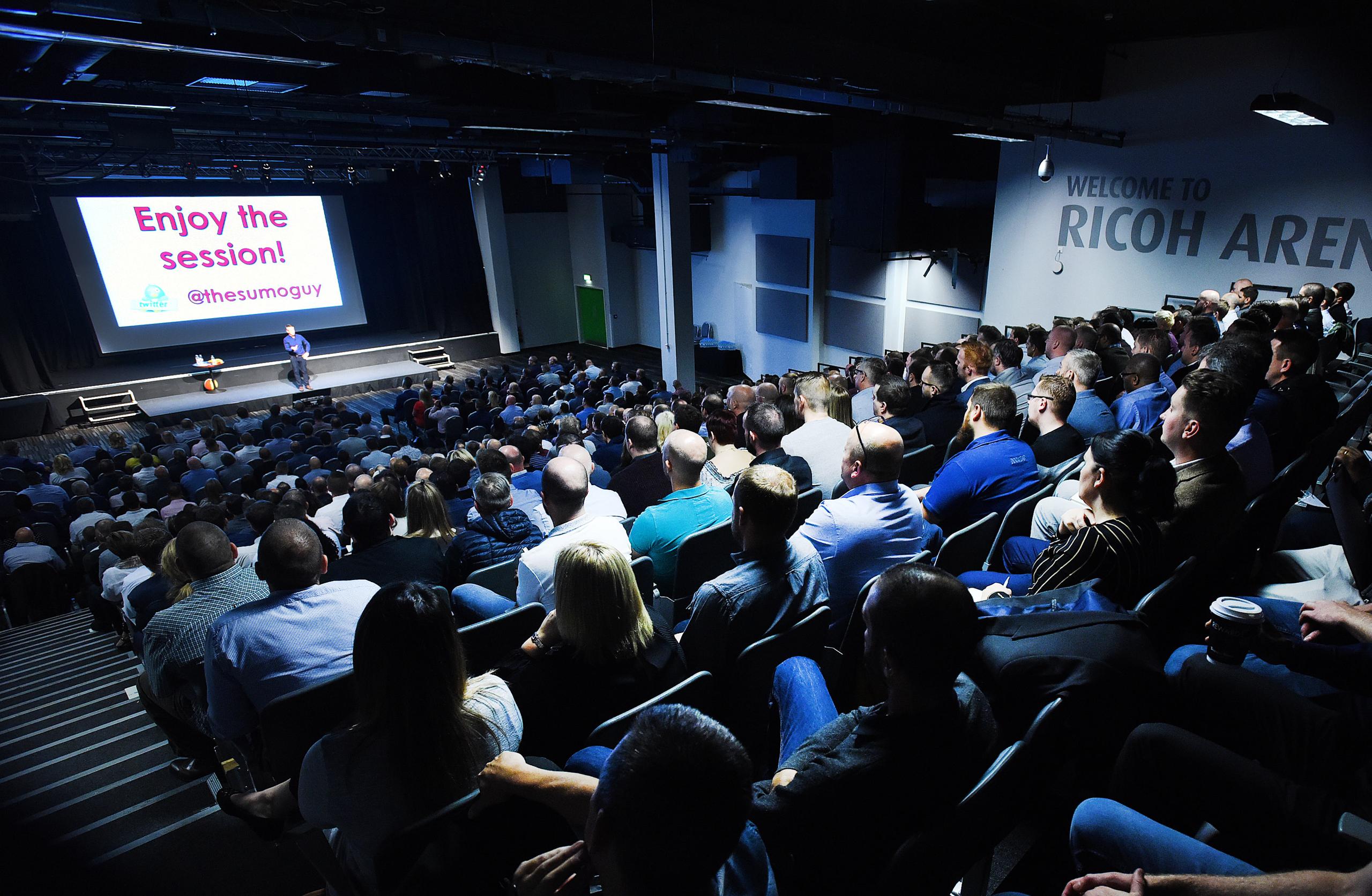 Paul McGee presenting to a packed auditorium at the Ricoh Arena, with “Enjoy the session!” on the screen